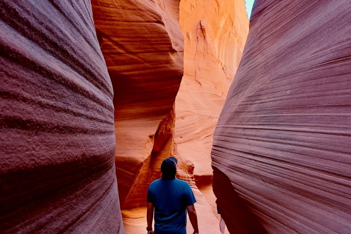 Person walking through narrow sandstone canyon with blue sky above.