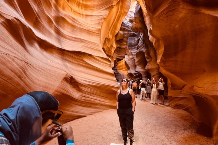 Tourist posing in narrow, winding sandstone canyon with textured orange-brown walls.